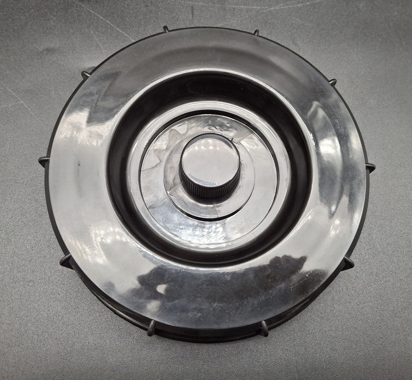 A circular plastic component with a ribbed central knob and small protruding tabs around the outer edge, photographed on a gray textured surface.