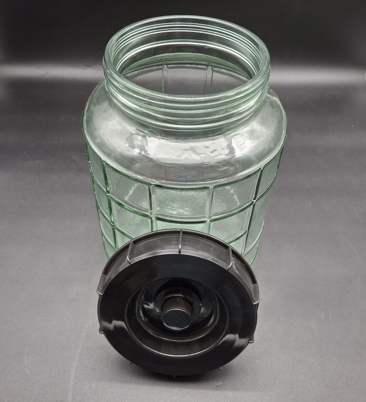 A clear ribbed glass jar with a threaded neck and a separate black screw-on lid, photographed on a dark gray background.