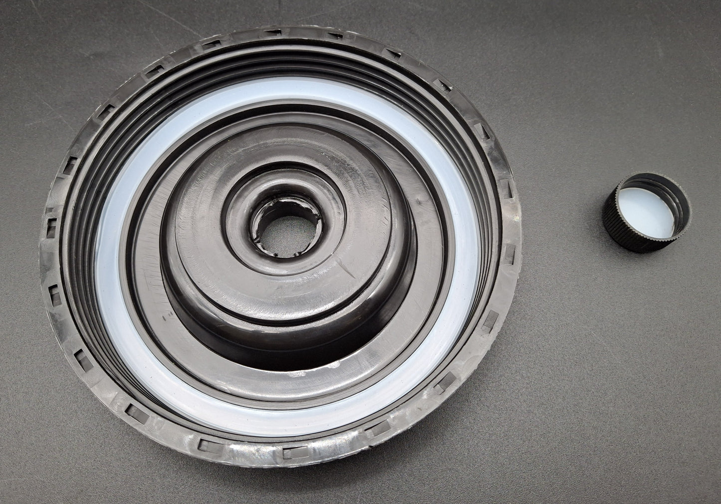 A circular black plastic bottle cap or lid component with a white inner ring, shown on a dark gray background with a smaller detached black cap piece to the right.