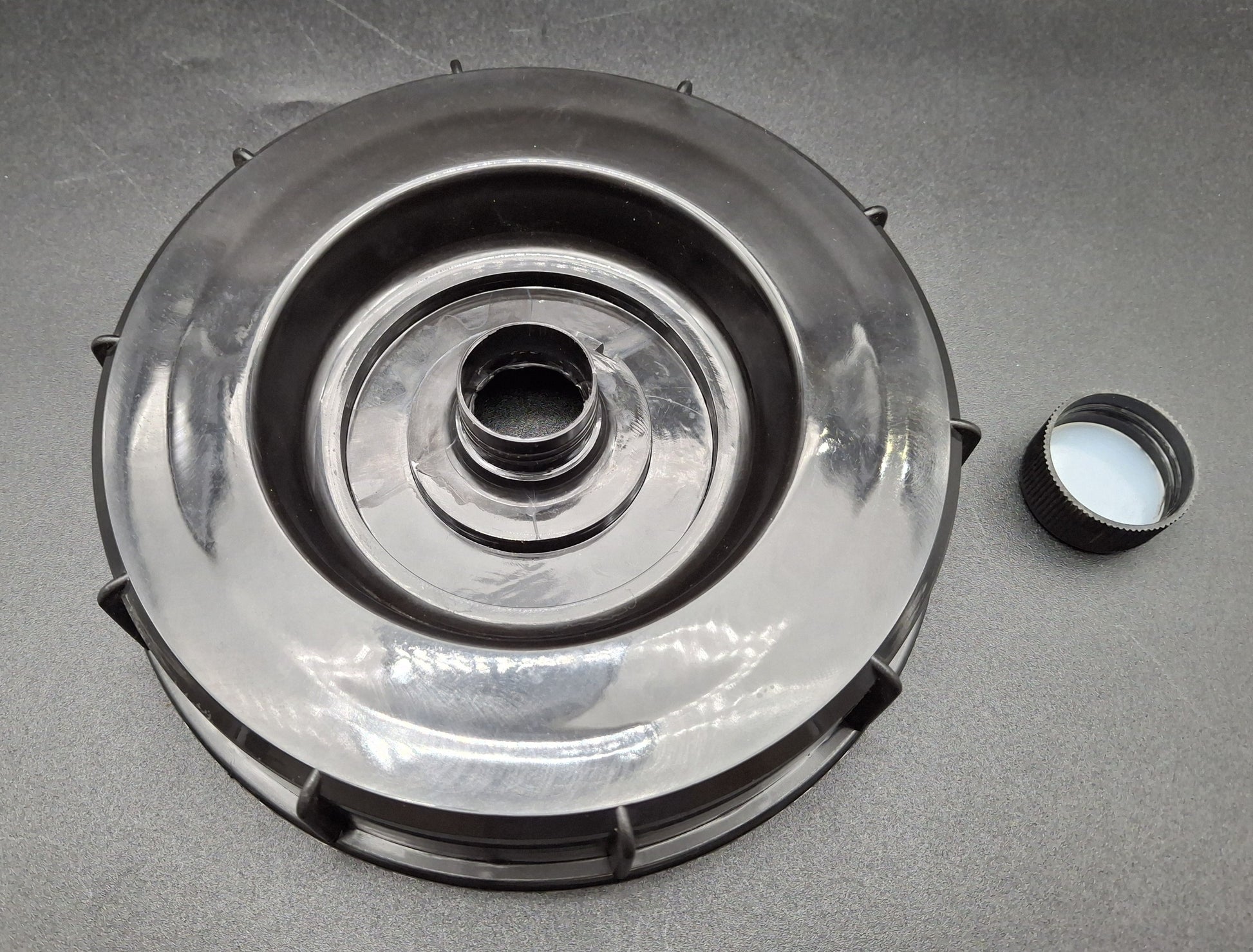 A large circular metallic mechanical part with a central hole, shown on a gray surface next to a small black cap with a white substance inside.