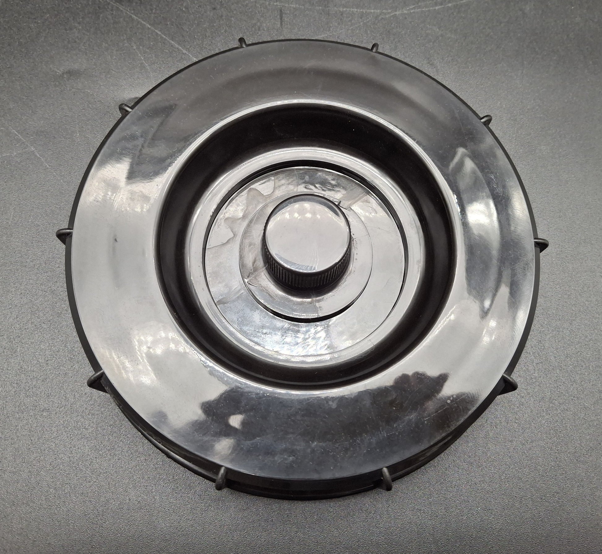 A circular plastic component with a ribbed central knob and small protruding tabs around the outer edge, photographed on a gray textured surface.