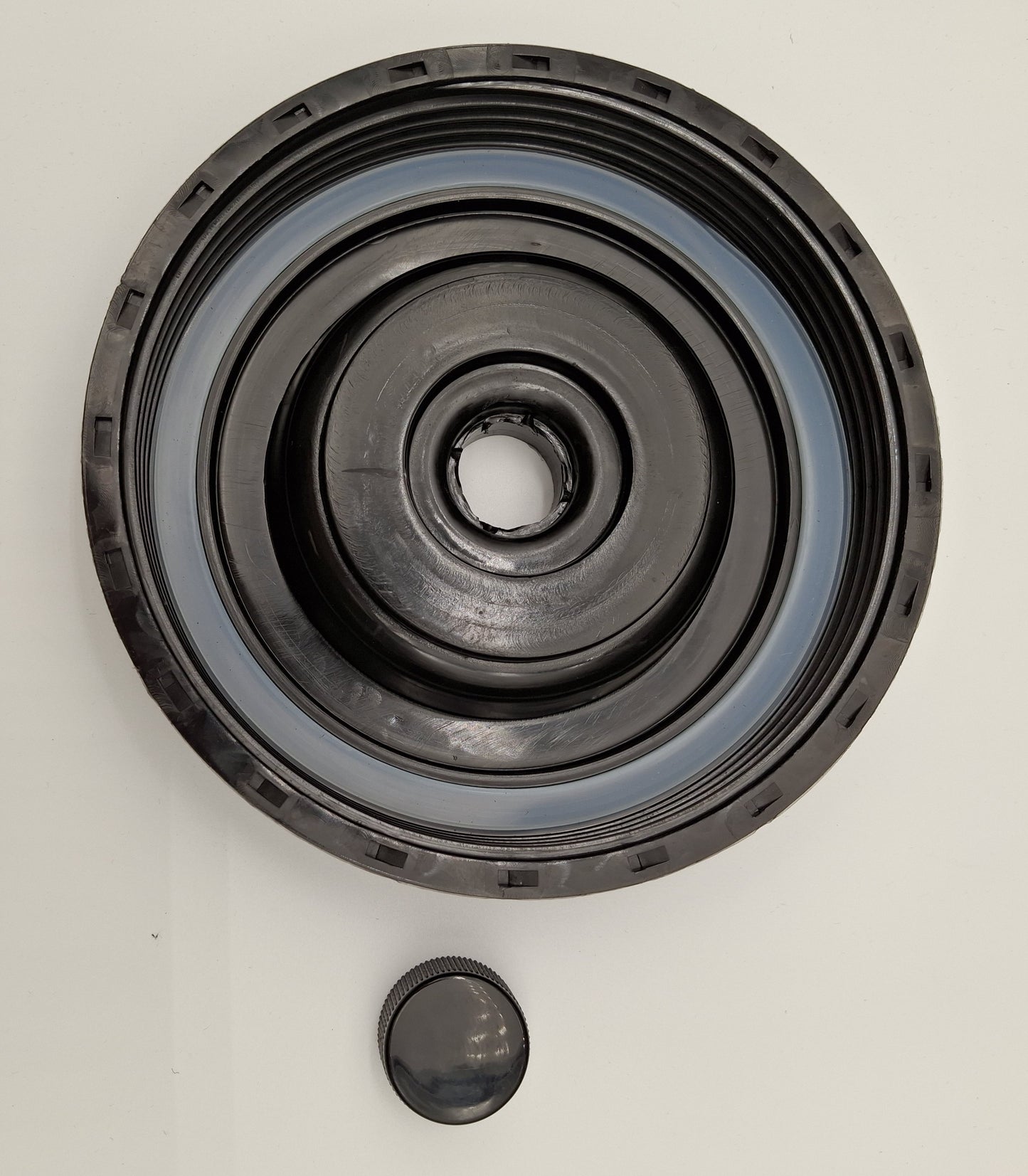 A black circular mechanical gasket or filter element with concentric grooves and a separate small matching cap below it, photographed on a plain light background.