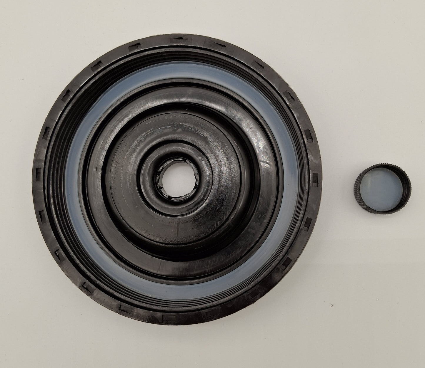 A black circular mechanical seal or bearing assembly with concentric rings, shown on a plain light background with a small matching cap or cover piece to the right.