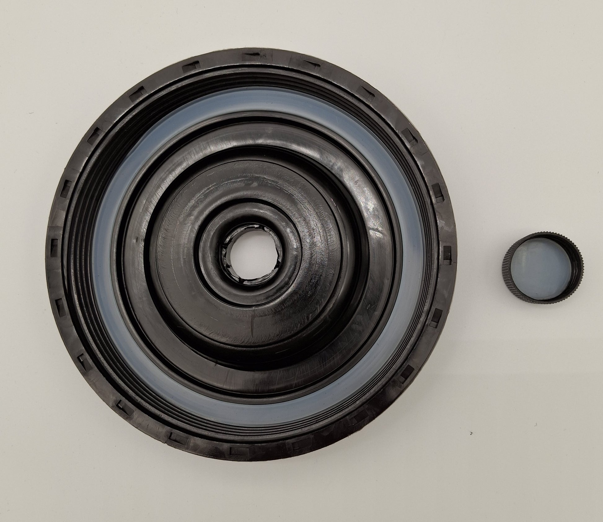 A black circular mechanical seal or bearing assembly with concentric rings, shown on a plain light background with a small matching cap or cover piece to the right.