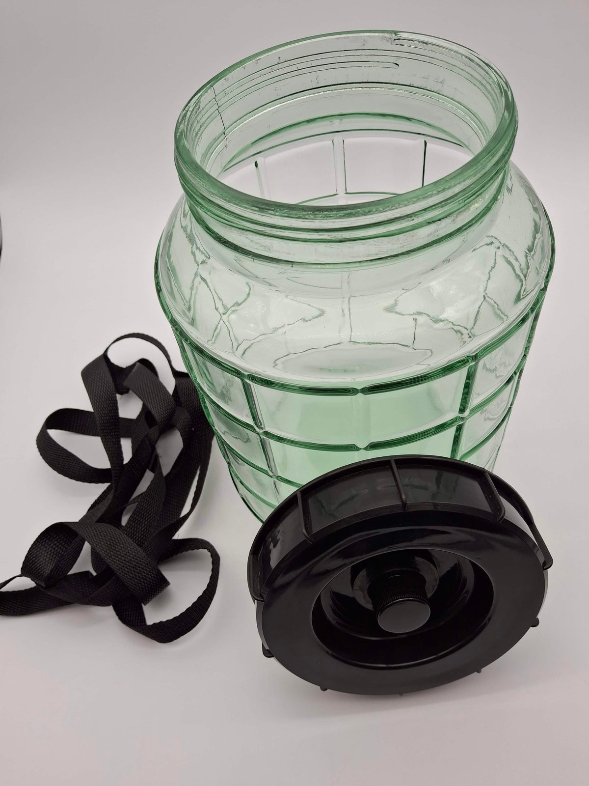 A clear green glass jar with a textured ribbed design, shown with a black plastic screw-on lid and a black carrying strap on a white background.