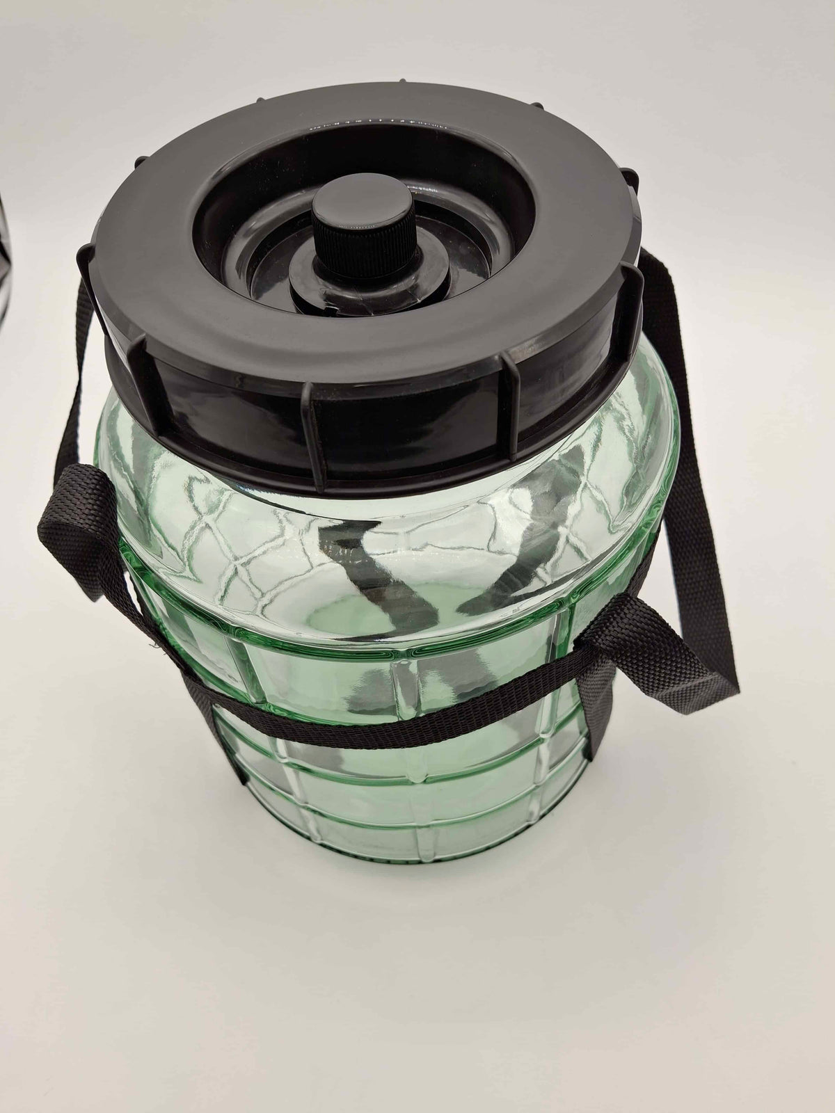 A clear plastic or glass jar with a black screw-on lid and black carrying straps, photographed on a plain light background.