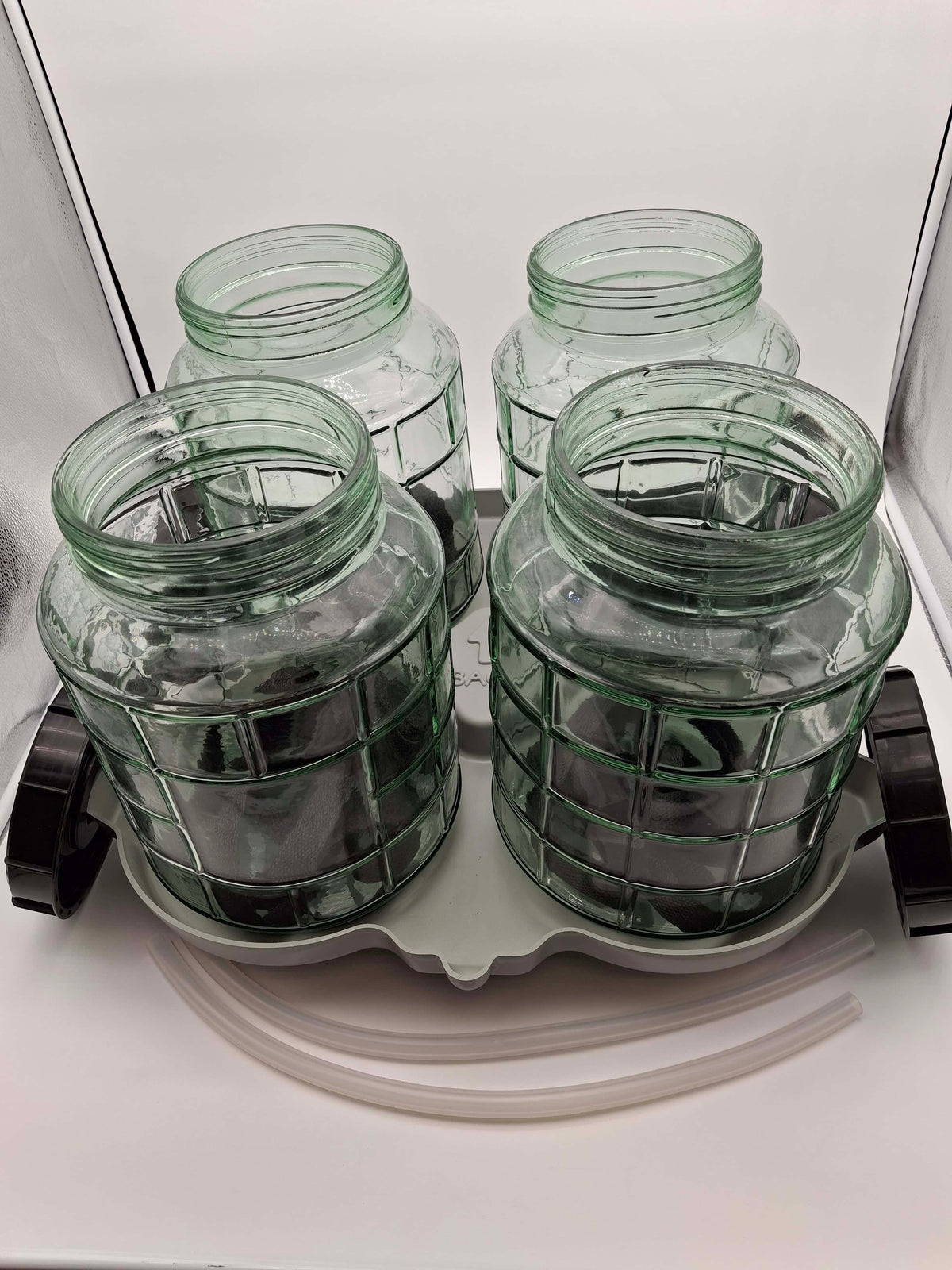 Four clear green glass jars with a grid pattern, arranged on a white plastic tray with two black caps and two clear tubes on a white background.