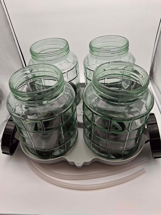 Four clear green glass jars with a grid pattern, arranged on a white plastic tray with two black caps and two clear tubes on a white background.