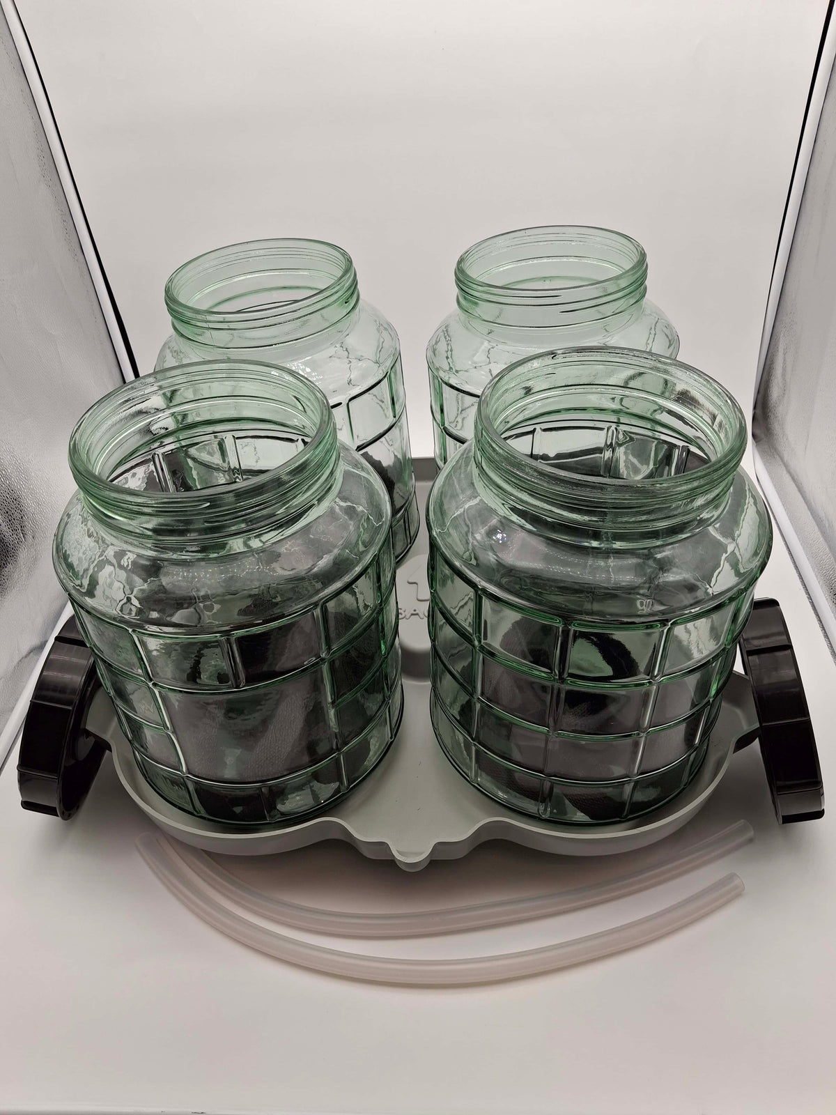 Four clear green glass jars arranged on a gray plastic tray with black side caps, plus two translucent tubes, photographed against a white studio background.