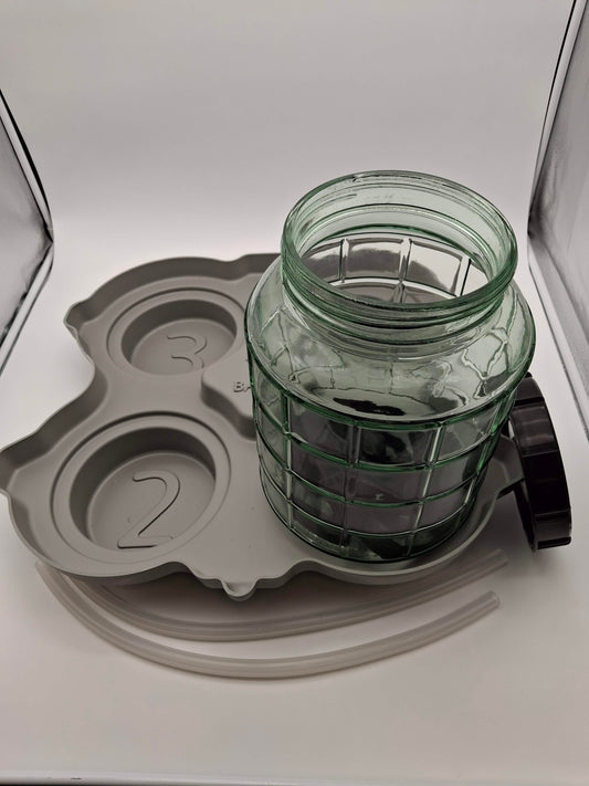 A clear ribbed glass jar with a dark green tint, shown inside a gray silicone mold tray with a black screw cap, on a white background.
