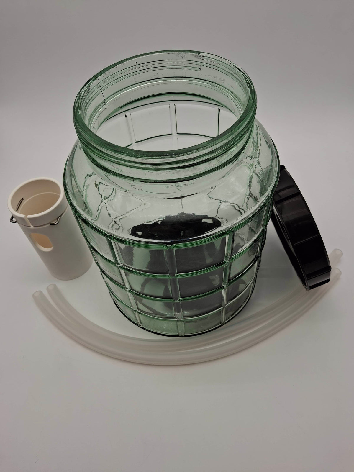 A large clear glass fermentation jar with a green tint, shown with its black lid, a white ceramic thermometer, and a translucent white plastic funnel on a white background.