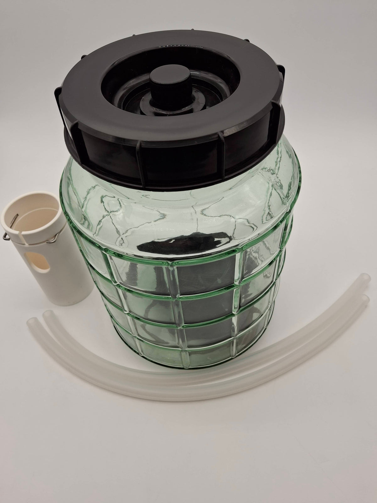 A clear glass fermentation or storage jar with a ribbed green-tinted body, a black screw-on lid, a white plastic funnel, and two curved clear plastic transfer tubes, photographed on a light gray studio background.