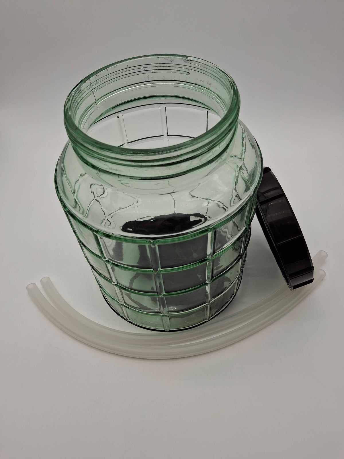 A clear green glass jar with a ribbed grid pattern, shown open with a black lid resting against it and two translucent white tubes curved in front, on a light gray background.