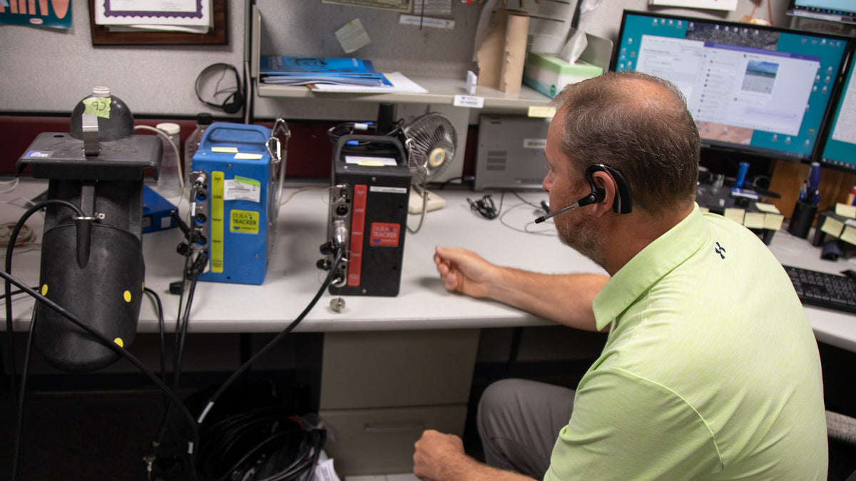 A man wearing a light green polo shirt and a headset sits at a desk in a technical office, looking at a device connected to a computer monitor.